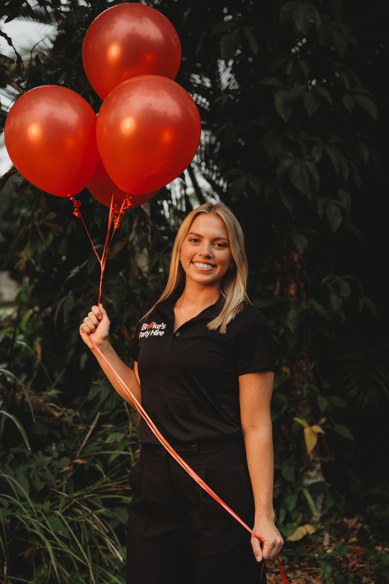 Woman holding red balloons outdoors with greenery in the background